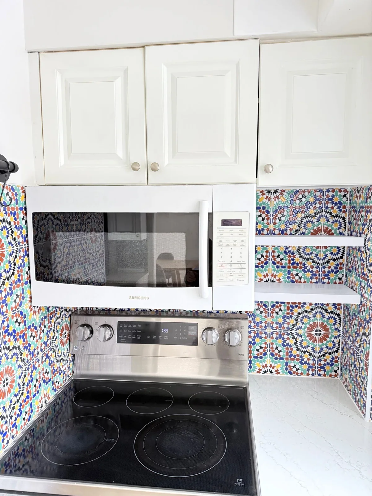 Small white shelves sit within a colorful geometric tiled corner beside a white microwave and cooker beneath white upper cabinets