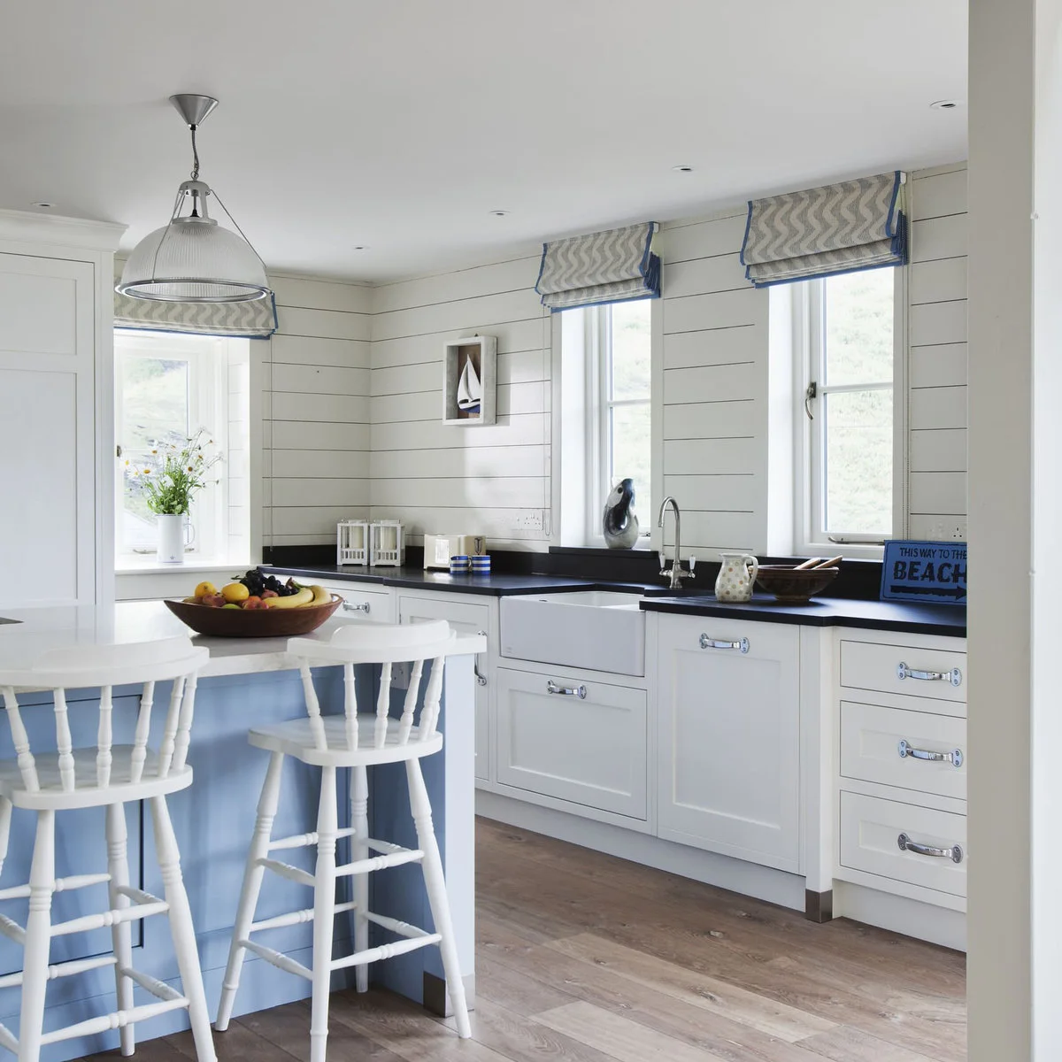Small framed sailboat picture on white boarded kitchen walls between windows with patterned blinds.