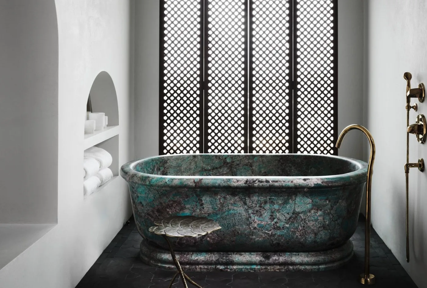 Mottled green freestanding bath beneath a dark lattice window with pale walls and a small stool