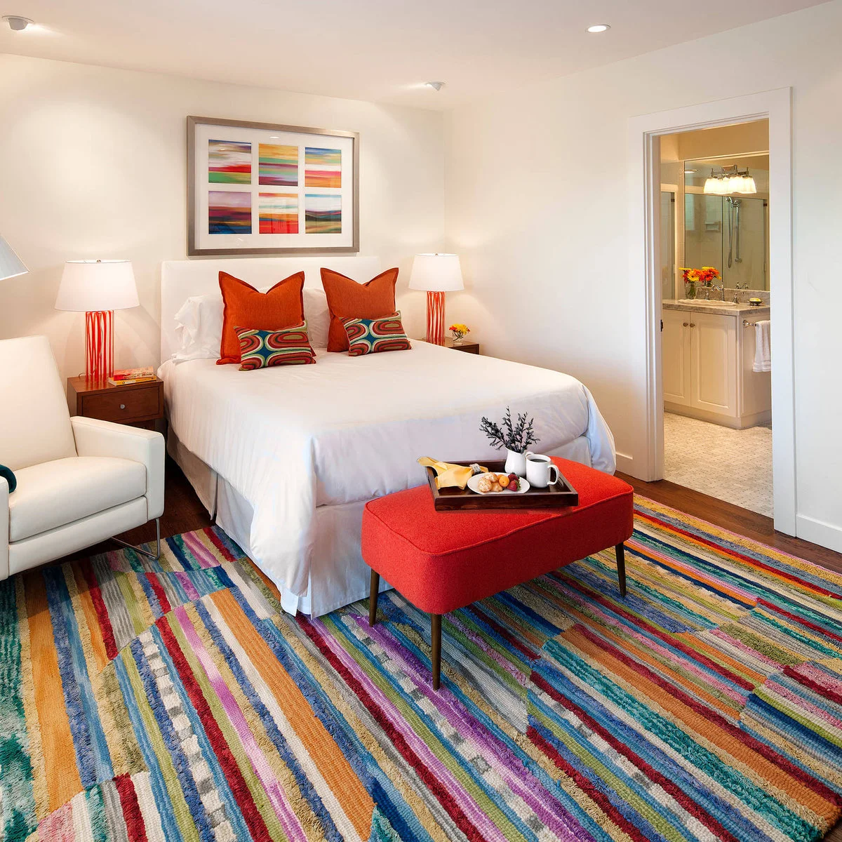 A multicolored striped rug surrounds a white bed and red-orange bench, with matching bright pillows and lamps.