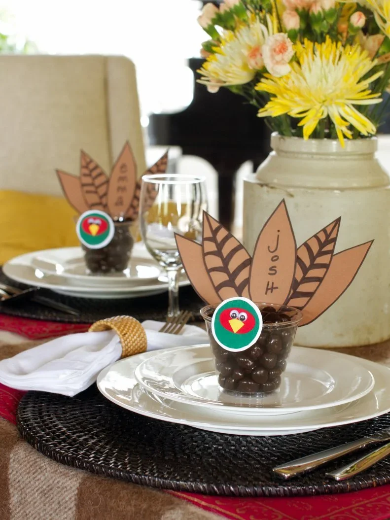 Paper turkey name cards rise from small clear cups on pale plates beside white napkins and simple glassware