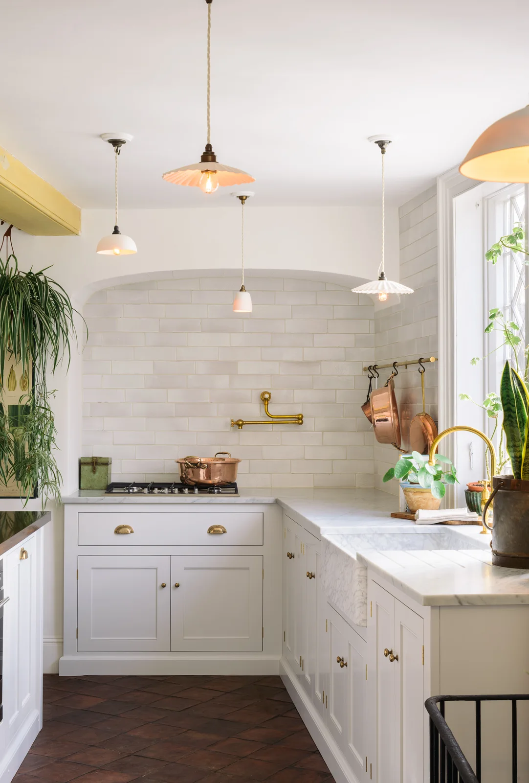 Dark terracotta floor tiles sit beneath white cabinets, hanging cookware and several pale pendant lights.