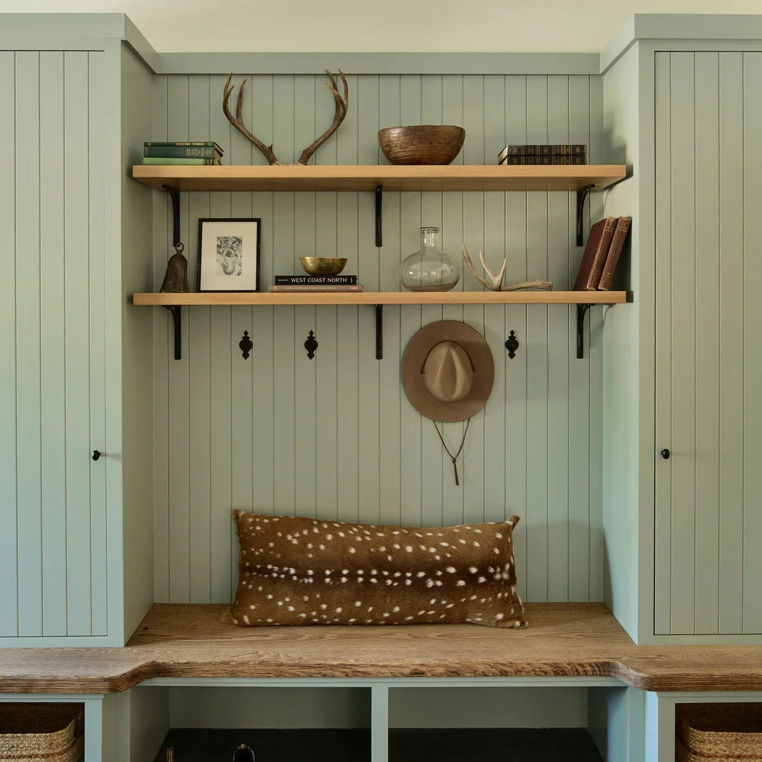 Muted green cupboards and vertical boards surround a wood bench, two wood shelves, dark hooks and woven storage baskets.