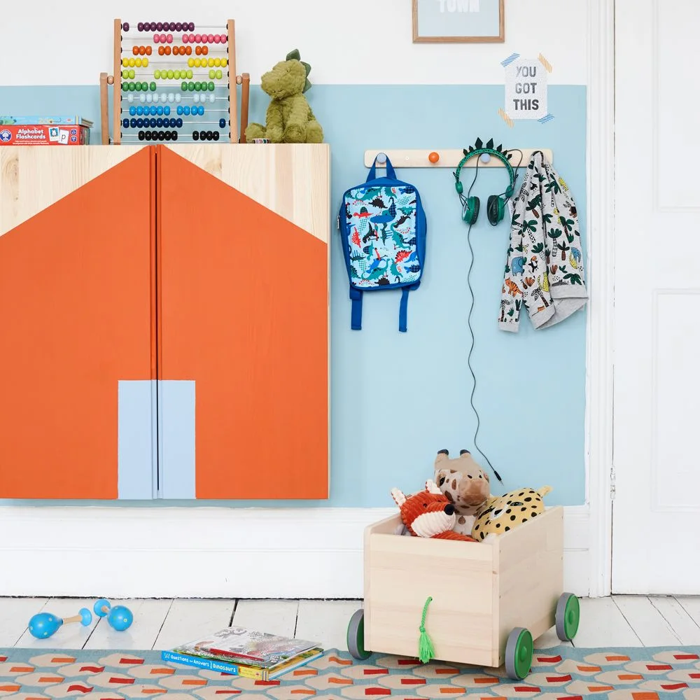 Wooden toy cart with green wheels holding soft animals below wall hooks and an orange cupboard