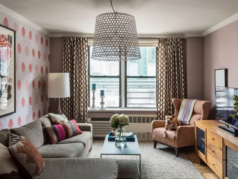 Gray sofa in a compact living room with pink patterned wallpaper, zigzag curtains and woven armchair.
