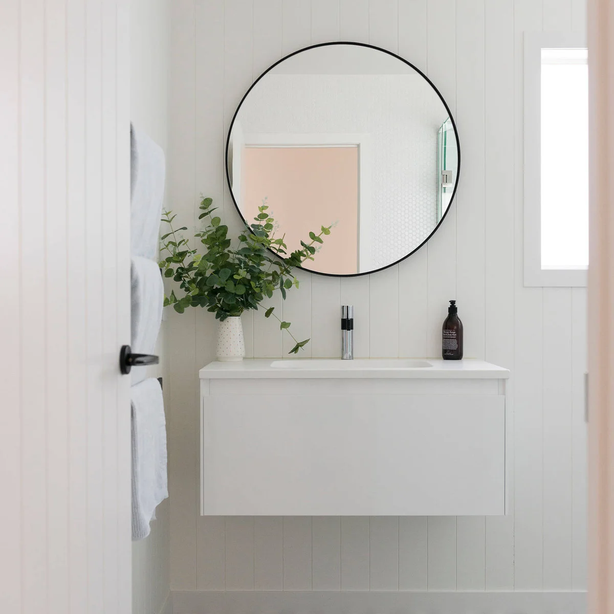 Small white wall-mounted vanity with a round dark-framed mirror against white vertical wall boards.