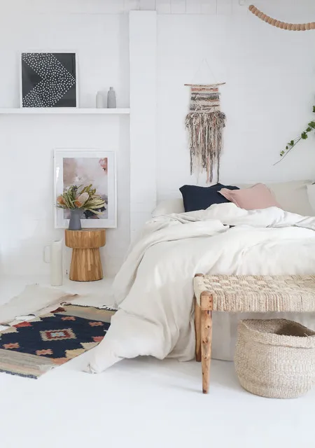 A pale bed sits beside a small wood table and fringed wall hanging with a wood bench, woven basket and darker patterned rug nearby