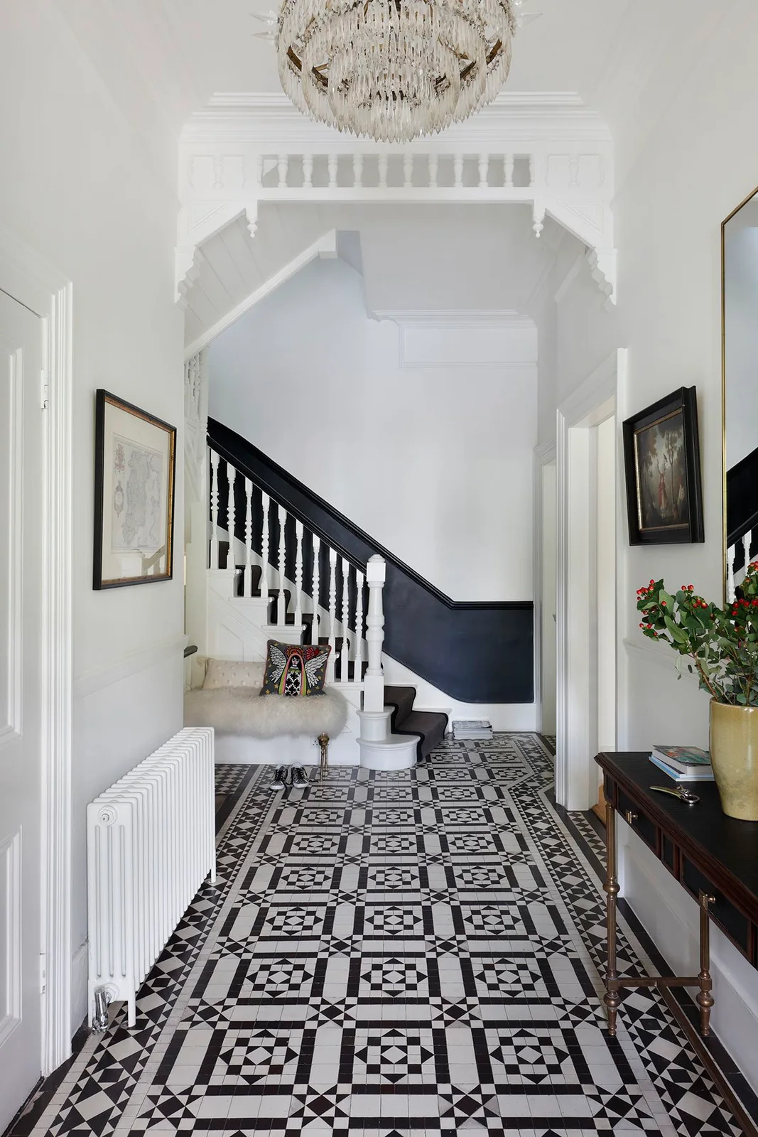 A white hallway has black-and-white patterned floor tiles below a dark lower stair wall and rail beside a pale seat and darker console