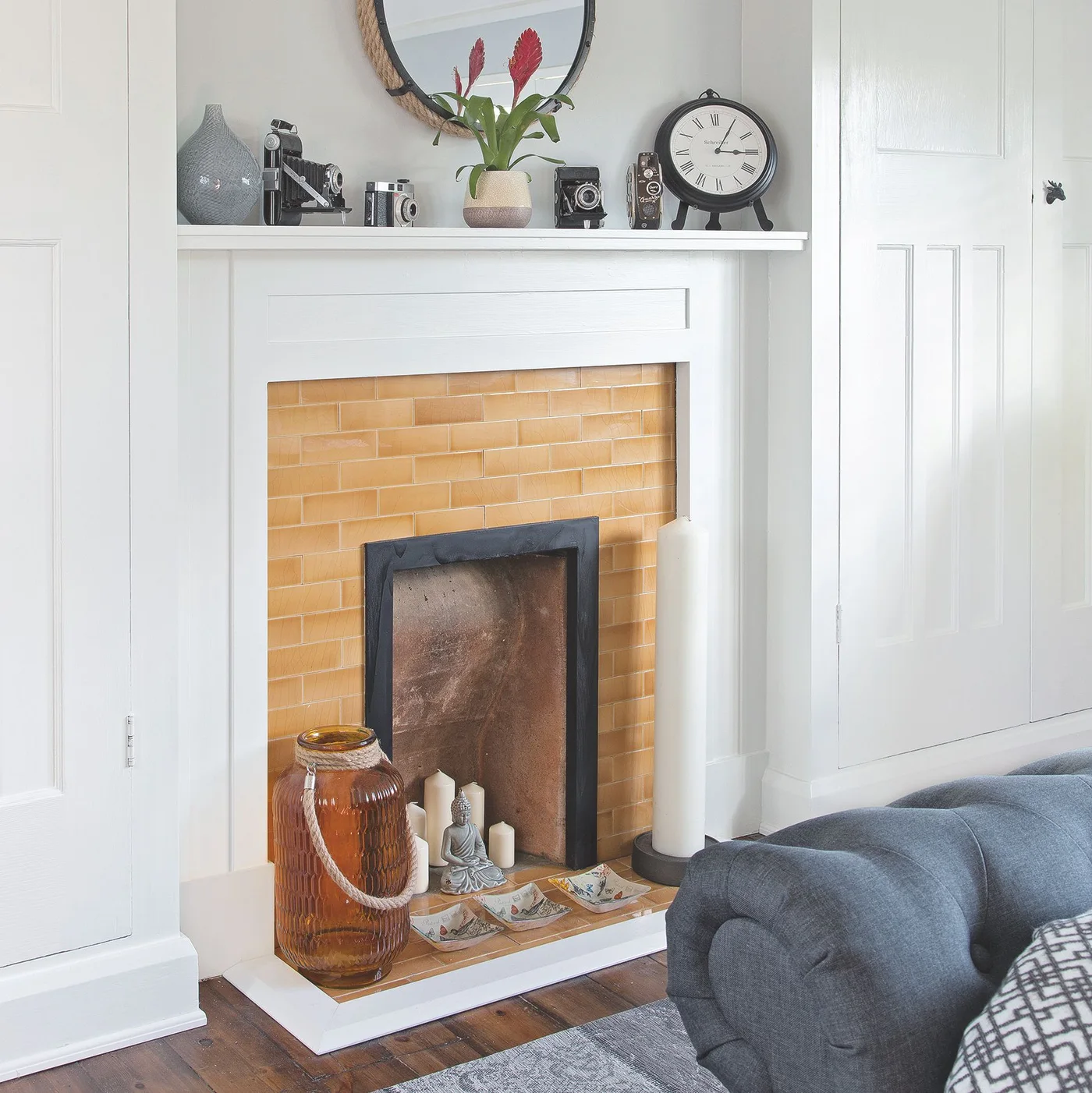 Amber rectangular tiles surround a closed fireplace opening and continue onto its hearth beside white cupboards and a gray sofa.