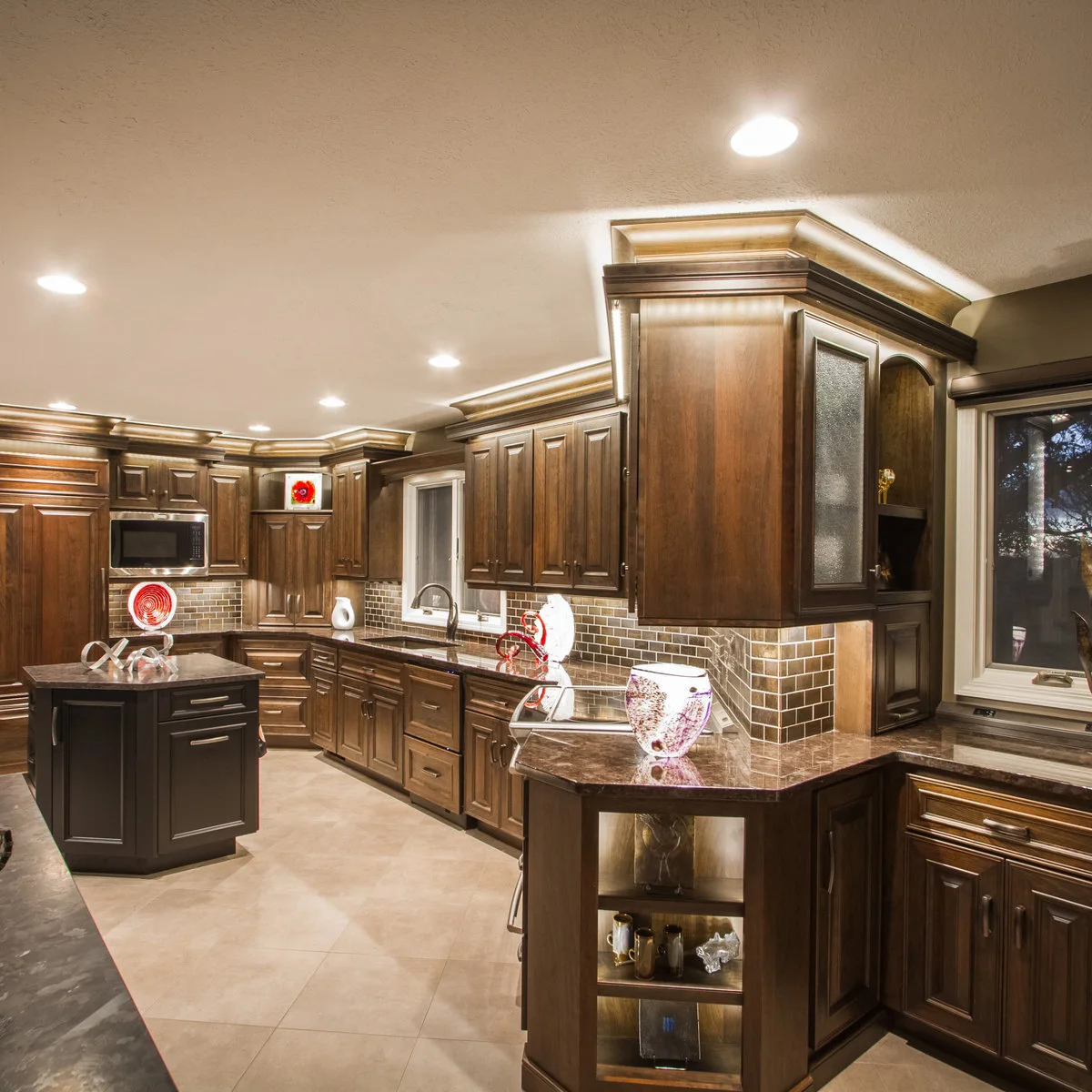 Dark wood cabinets have light above the crown, beneath the cupboards and inside small display shelves, with round ceiling lights overhead.