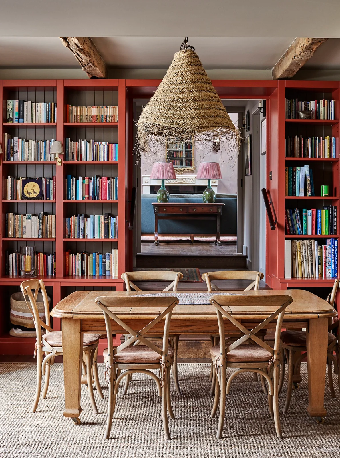 Red shelves filled with books frame a doorway behind a wooden table and cross-back chairs beneath a woven pendant