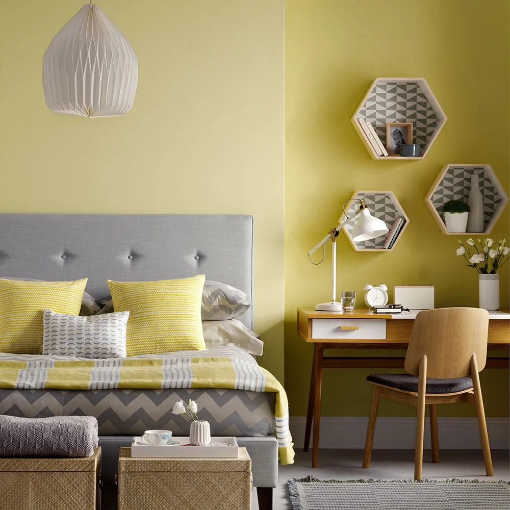 Pale yellow walls surround a gray upholstered bed beside a desk with geometric wall boxes, a wood chair and a white pendant.