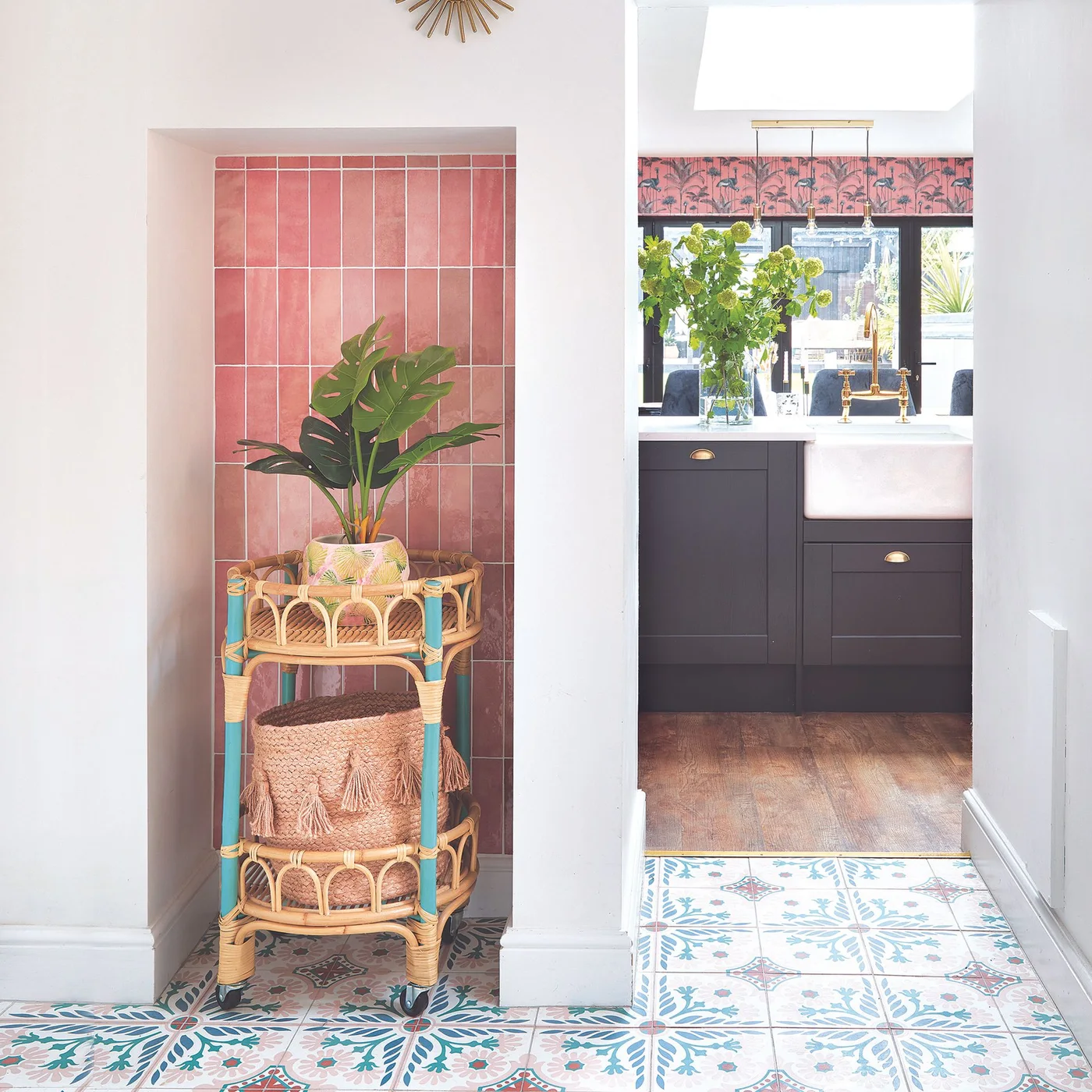 Pink rectangular tiles line a narrow alcove beside a kitchen sink and patterned floor, with a woven plant stand in front