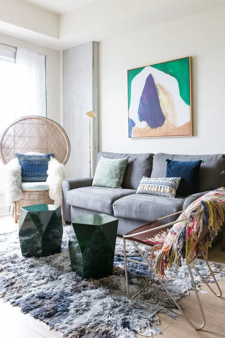 A gray sofa sits beneath green, purple and pale abstract artwork, with a woven chair, metal-framed accent chair and two dark green faceted tables.