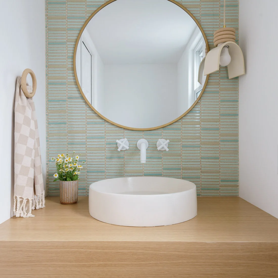 Fine pale green horizontal tiles rise behind a round warm-metal mirror, white wall taps, a round vessel basin and a wood counter with a sculptural pendant.