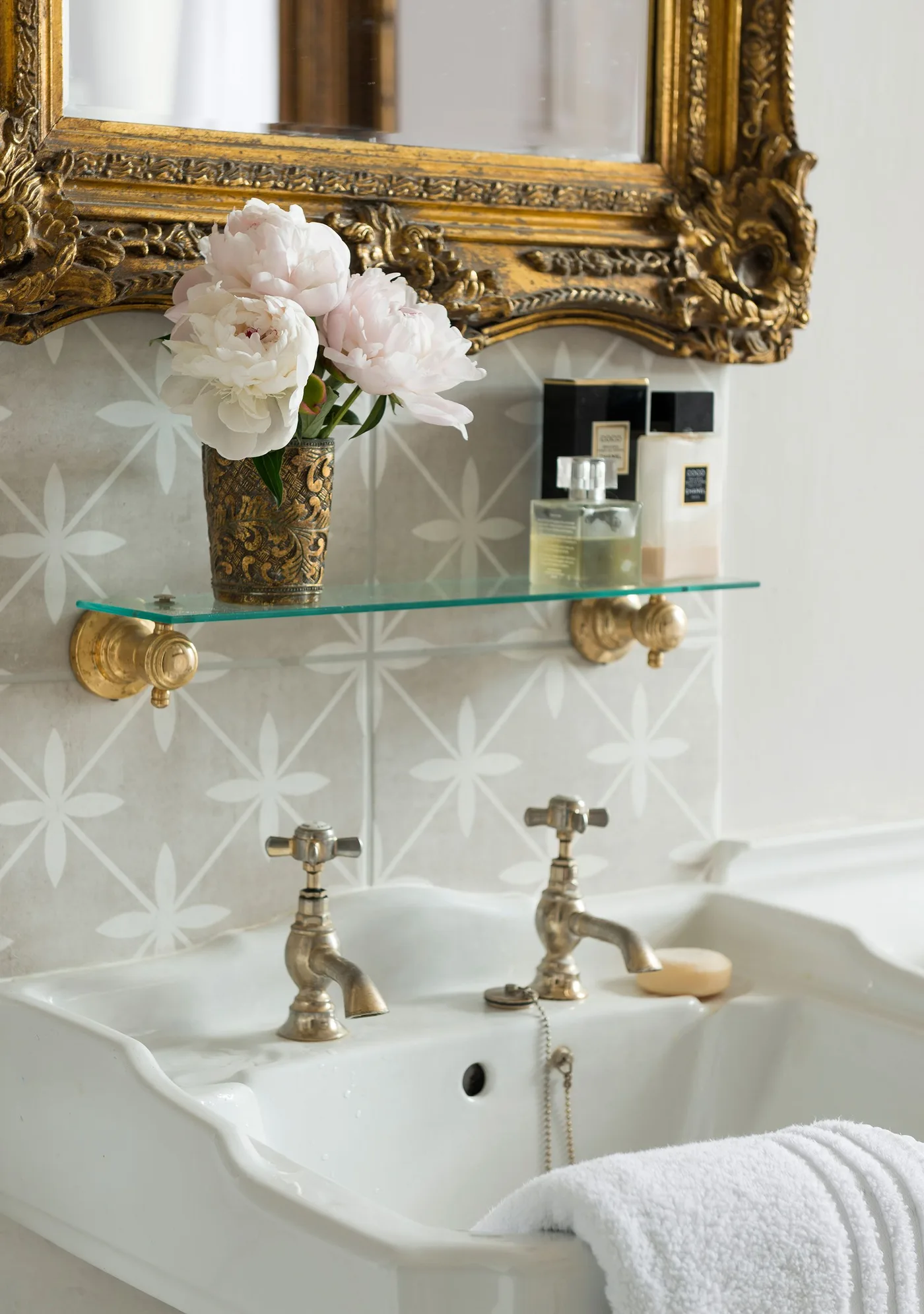 Ornate gold-toned mirror above a white basin, glass shelf, patterned wall and traditional-style taps.