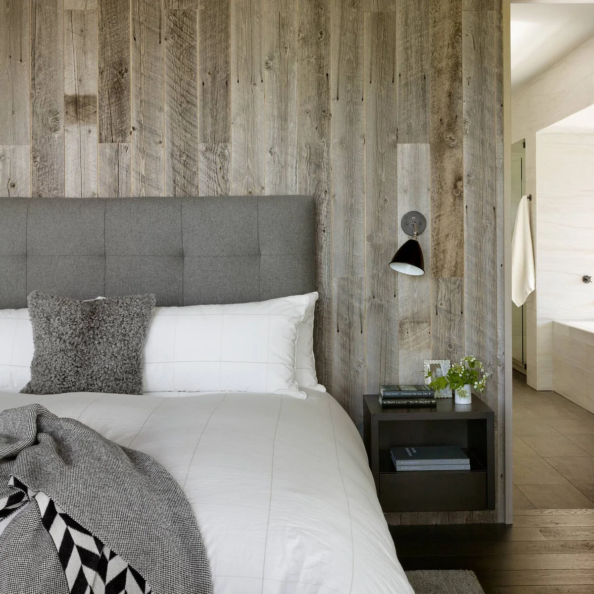 A small dark tilted conical wall shade sits above a floating dark nightstand beside a gray bed and vertical wood boards.