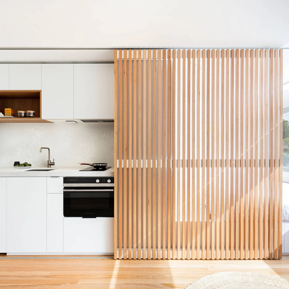 A tall wood slat screen covers part of a compact white kitchen with a sink and built-in oven.