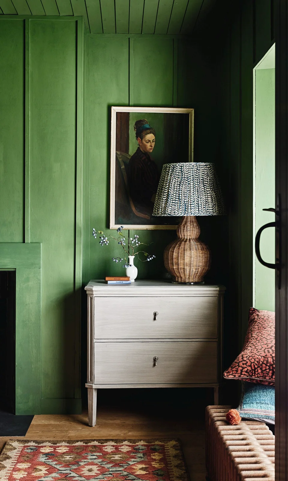 Bright green wall and ceiling boards surround a pale two-drawer cabinet with a woven lamp and framed portrait