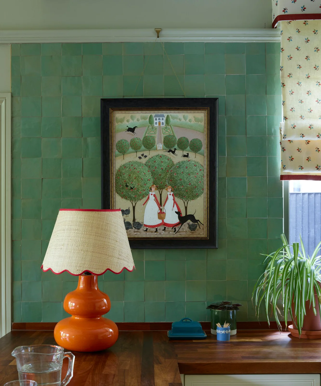 Green square wall tiles sit behind a framed picture and orange lamp beside a wooden counter and floral window fabric