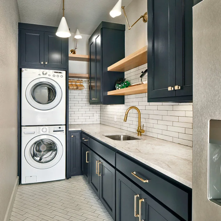 Navy cabinets surround stacked white machines beside a wood counter, utility sink, white brick-pattern backsplash and wood shelves.