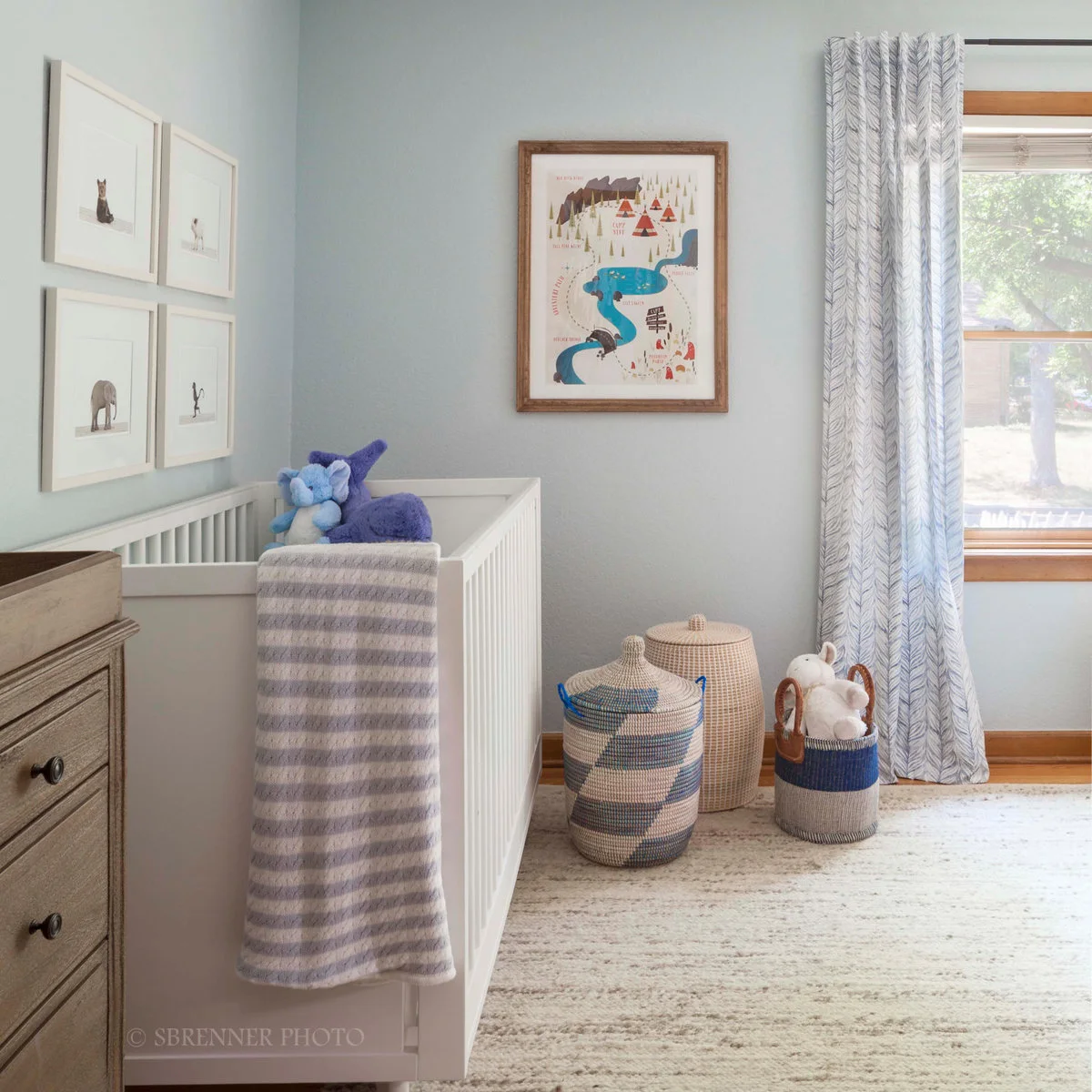 Pale blue nursery with a white crib, animal pictures and three woven baskets beside patterned curtains