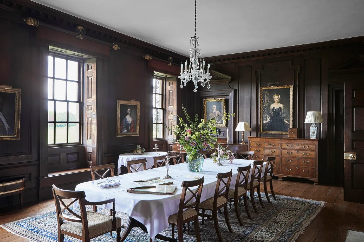 Full-height dark dining wall panels with portraits, tall windows, a white-clothed table and wood chairs