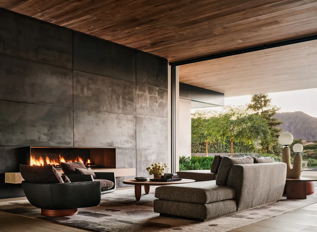 Textured gray seating sits beside a broad dark-paneled fireplace wall and a glazed garden opening.