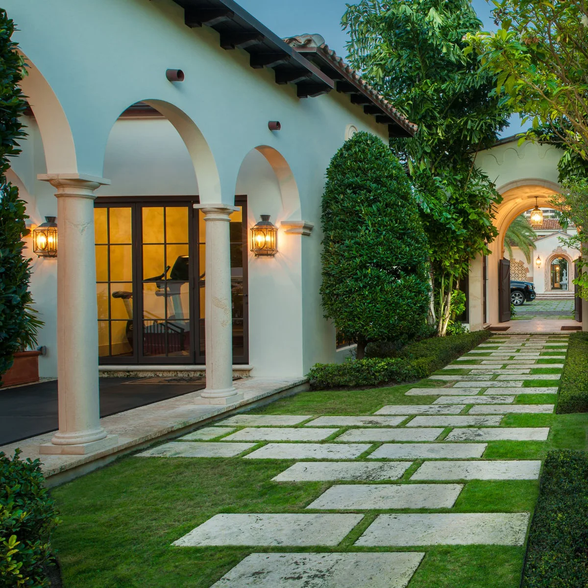 An arched cream house beside a path of pale rectangular slabs with green joints, clipped shrubs and taller leafy trees.