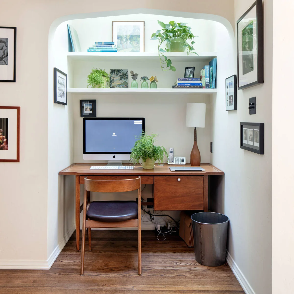 Compact wood writing desk beneath an arched alcove with white shelves, a monitor and matching wood chair