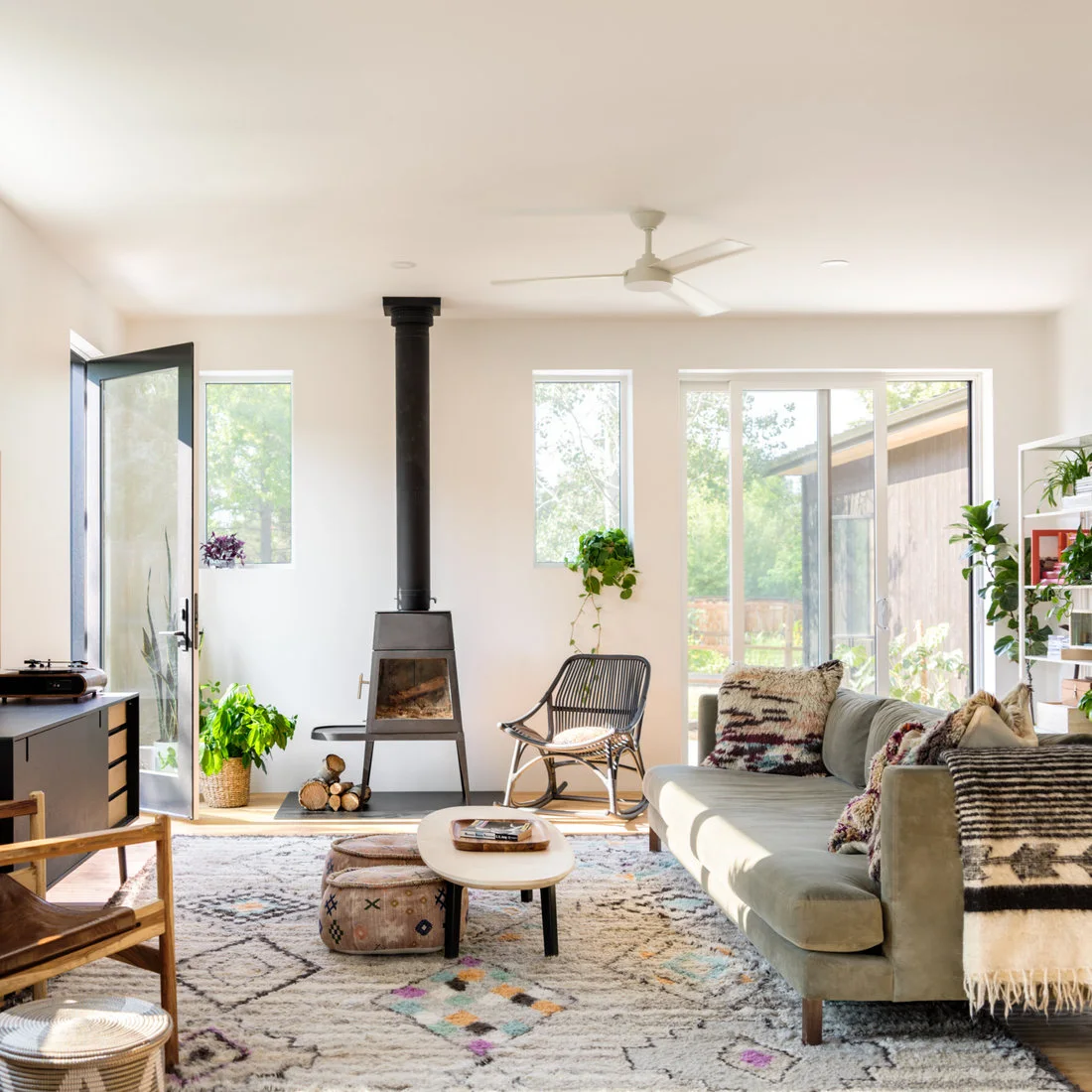 A bright living room has a dark tapered stove between windows, a pale green sofa, wood seating, a small rounded coffee table and a lightly patterned rug.