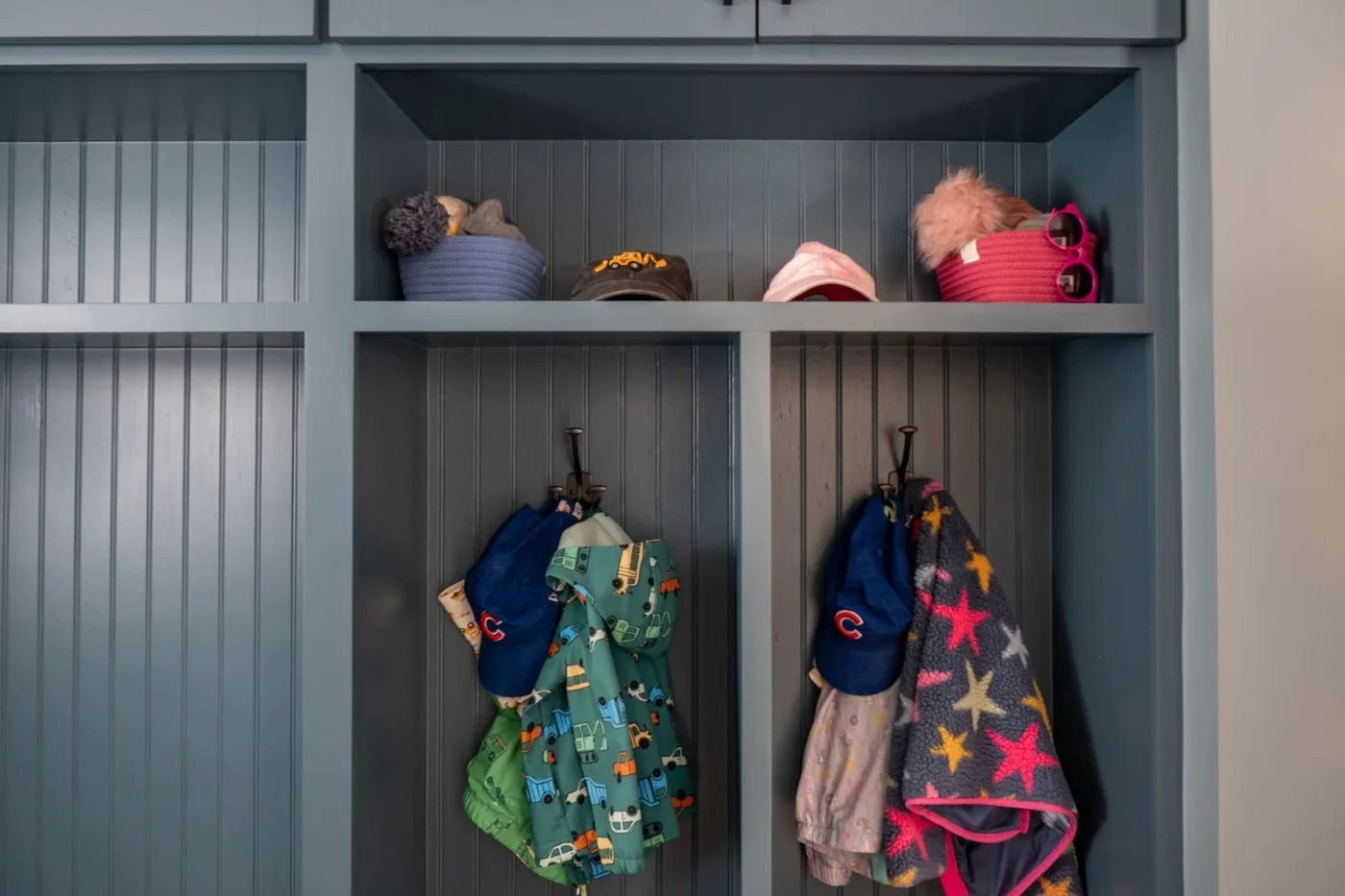 Blue divided cubbies holding baskets and hats above hooks with colorful children’s backpacks and jackets.