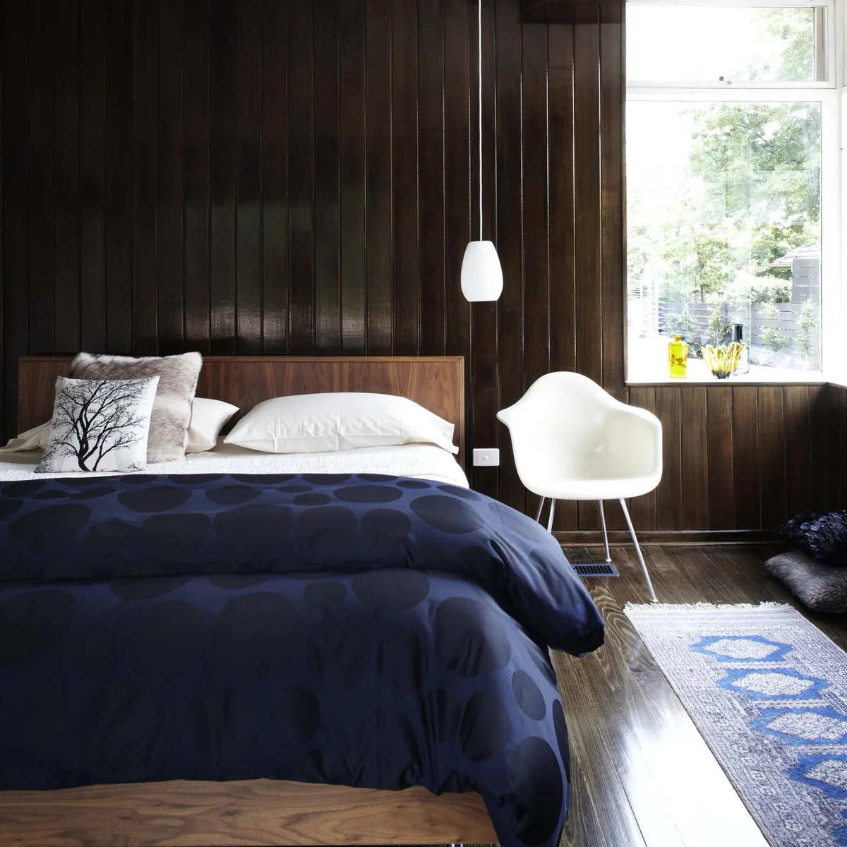 Dark brown paneled bedroom wall with a wooden headboard, blue bedding and a white chair.