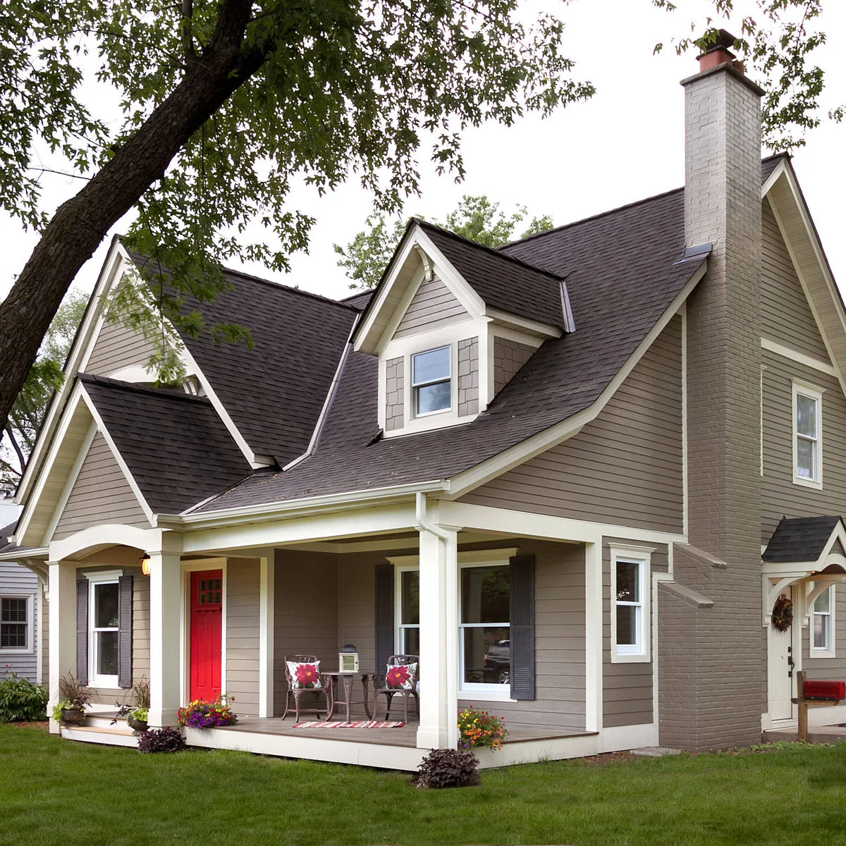 A gray Cape-style house with dormers has a red front door, white porch posts and two small chairs beside the entry.