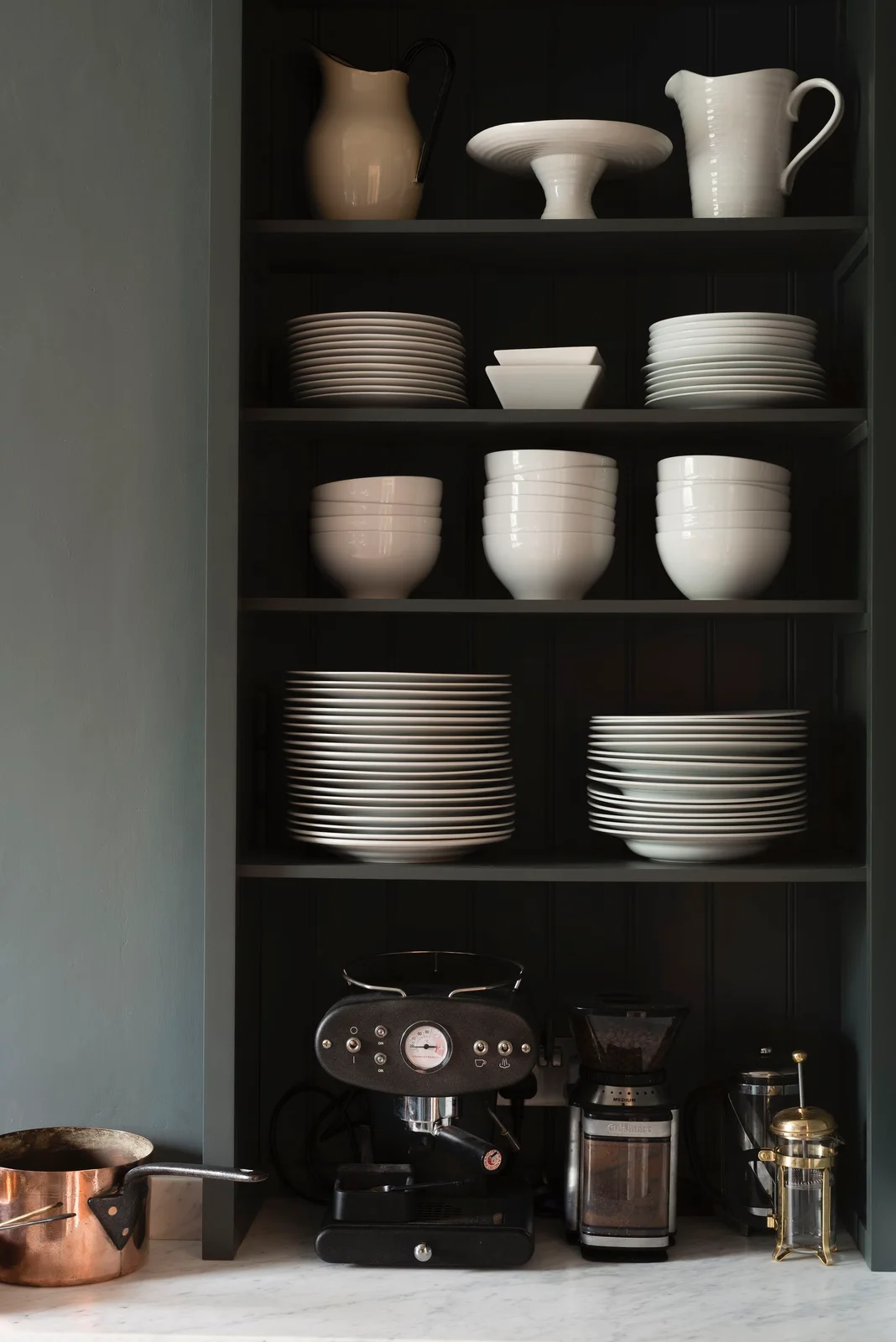 Dark countertop cupboard with stacks of pale dishes above a coffee machine and grinder