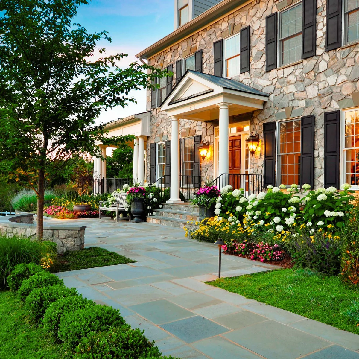 A wide pale path leads to a white portico on a stone house between beds of white, pink and yellow flowers.
