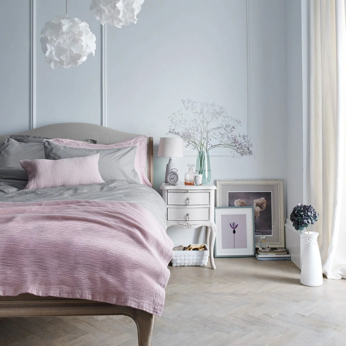 Dusty pink throw and cushions on a gray bed beside a small white bedside chest