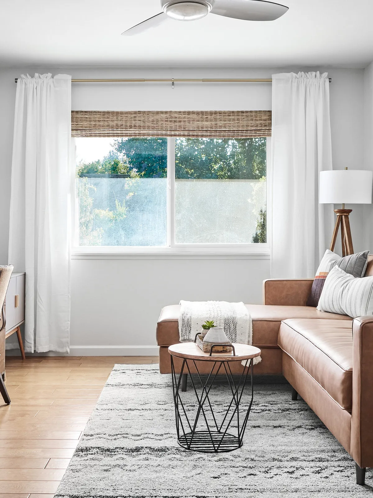 A tan sofa sits beside a broad window with a woven blind and pale curtains.