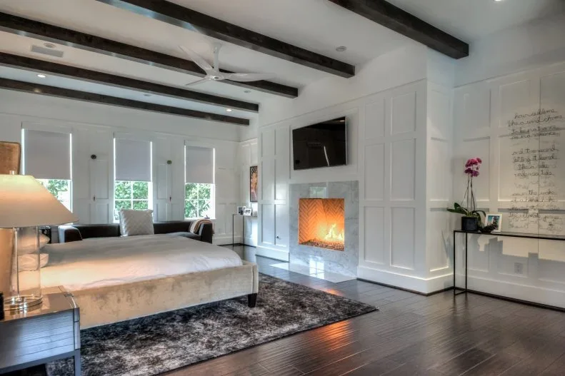 A bedroom with dark ceiling beams has a pale fireplace opening beside white paneled cupboards, a bed and a narrow console table