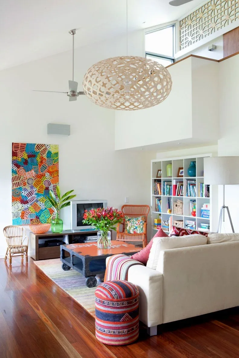 Tall white living room with a woven pendant, colorful painting, bookcase and striped cushions.