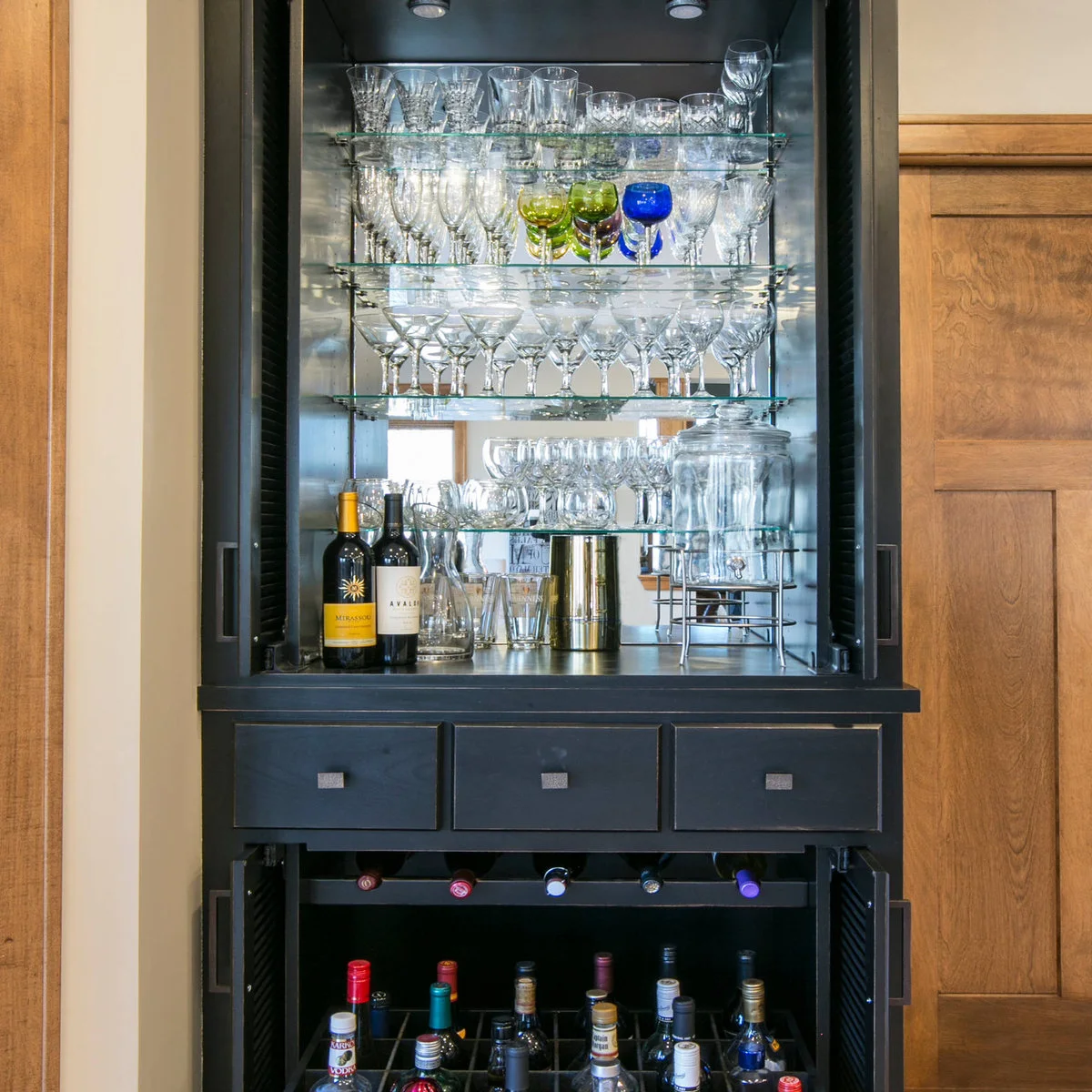 Open dark drinks cabinet with glass shelves, mirrored backing, stemware, bottles and shallow drawers