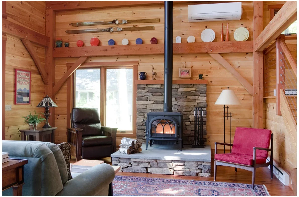 A black stove on a raised stone-faced hearth sits in a timber-lined living room with a dark armchair, red accent chair and patterned rug.
