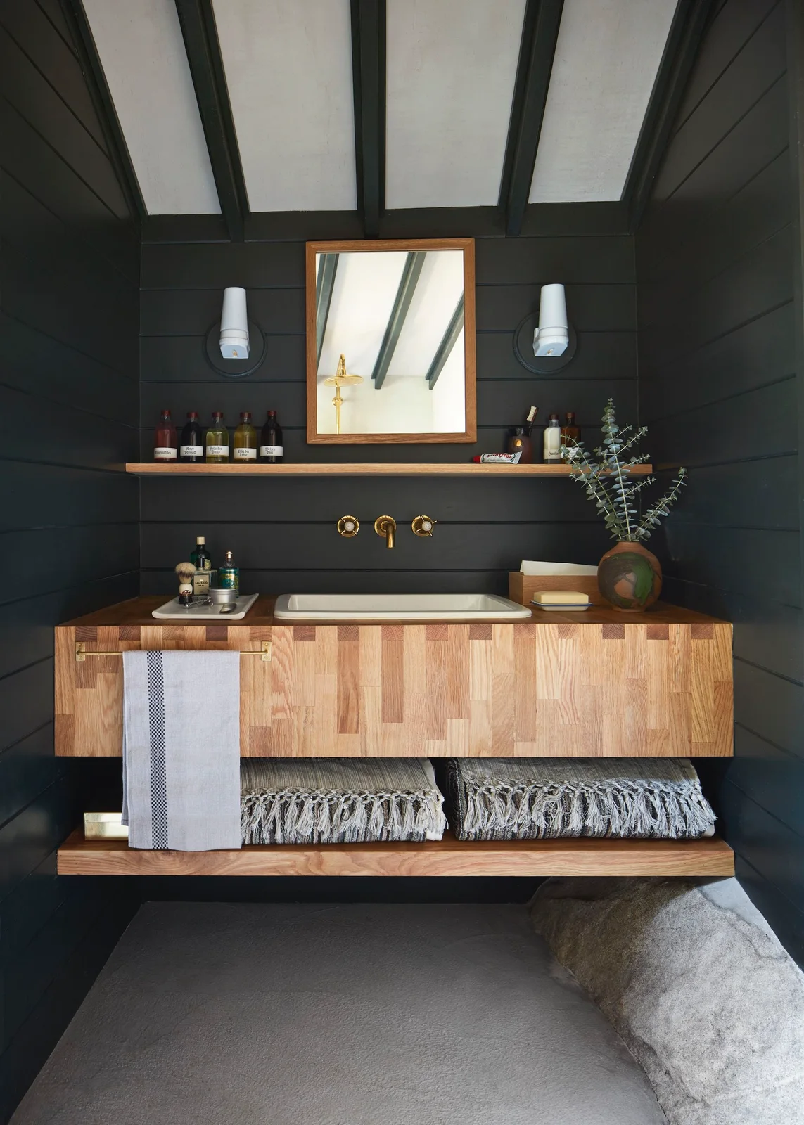 A wood vanity with an open lower shelf and pale inset basin sits beneath a rectangular mirror beside dark horizontal boards, a narrow shelf and two white wall lights