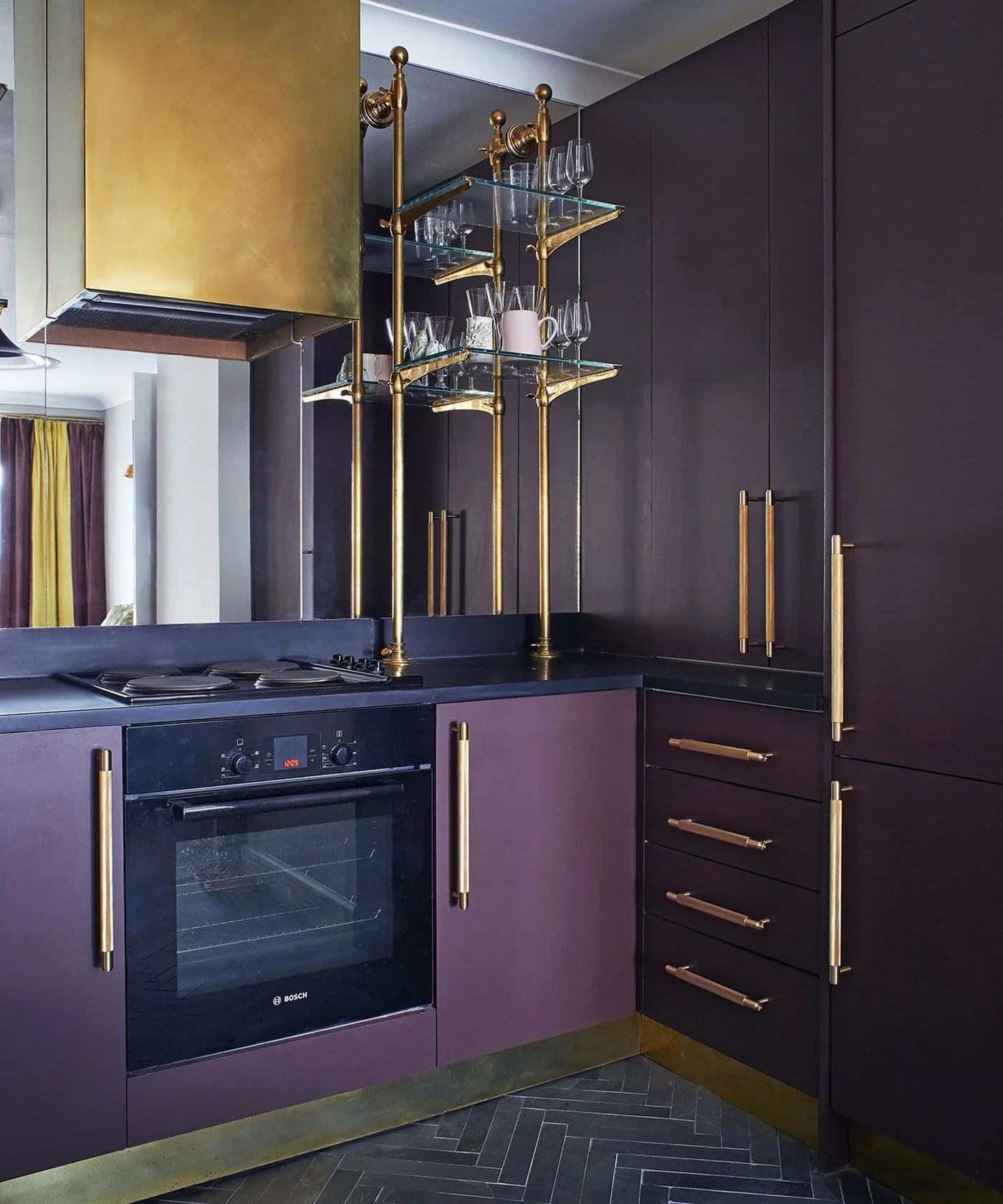 Kitchen with deep plum flat-front cabinets, long gold-colored handles, dark counter, warm metal hood, suspended glass shelves, and dark herringbone floor.