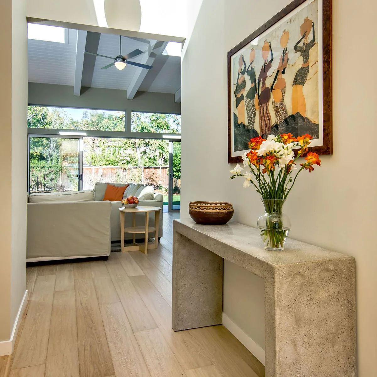 A pale solid console beneath framed art with a clear vase of orange-and-white flowers and a patterned bowl.
