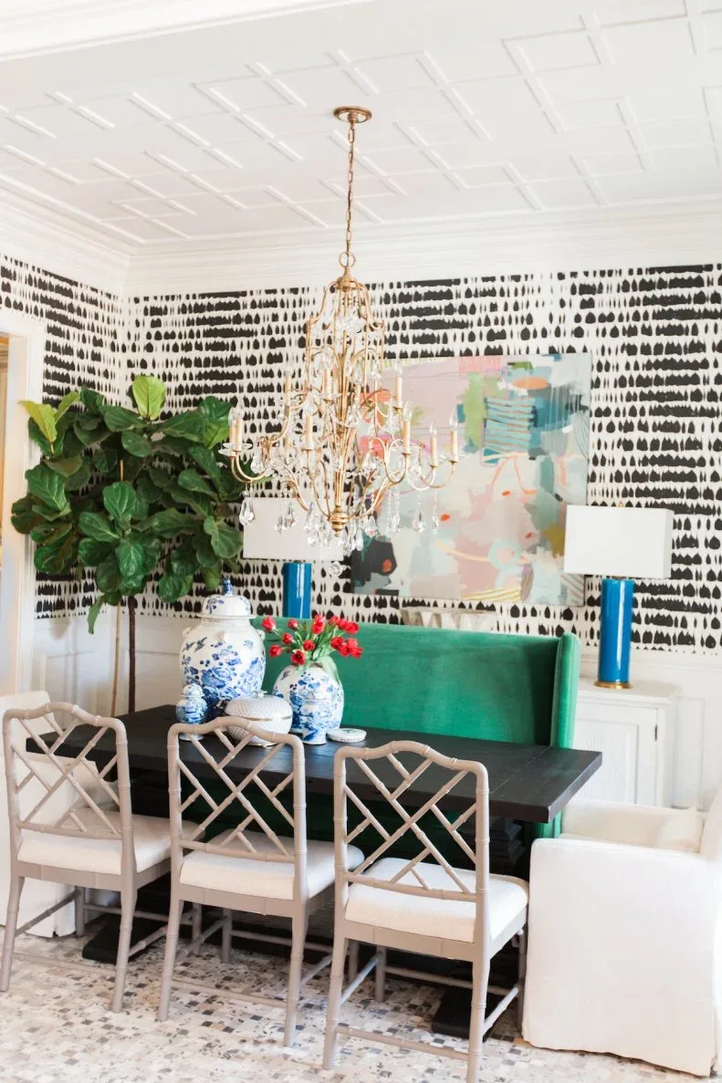 Dining room with black-and-white wallpaper, green banquette, pale open-backed chairs, dark table and gold-colored chandelier