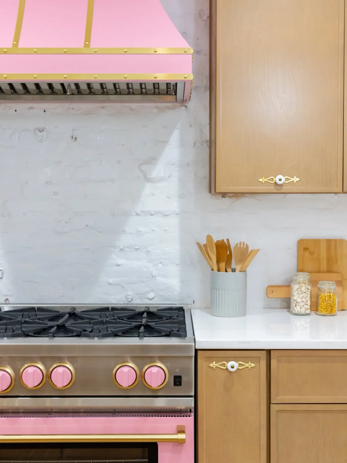 White oak cabinet doors with decorative pulls sit beside a pink range and matching hood against a pale textured backsplash.