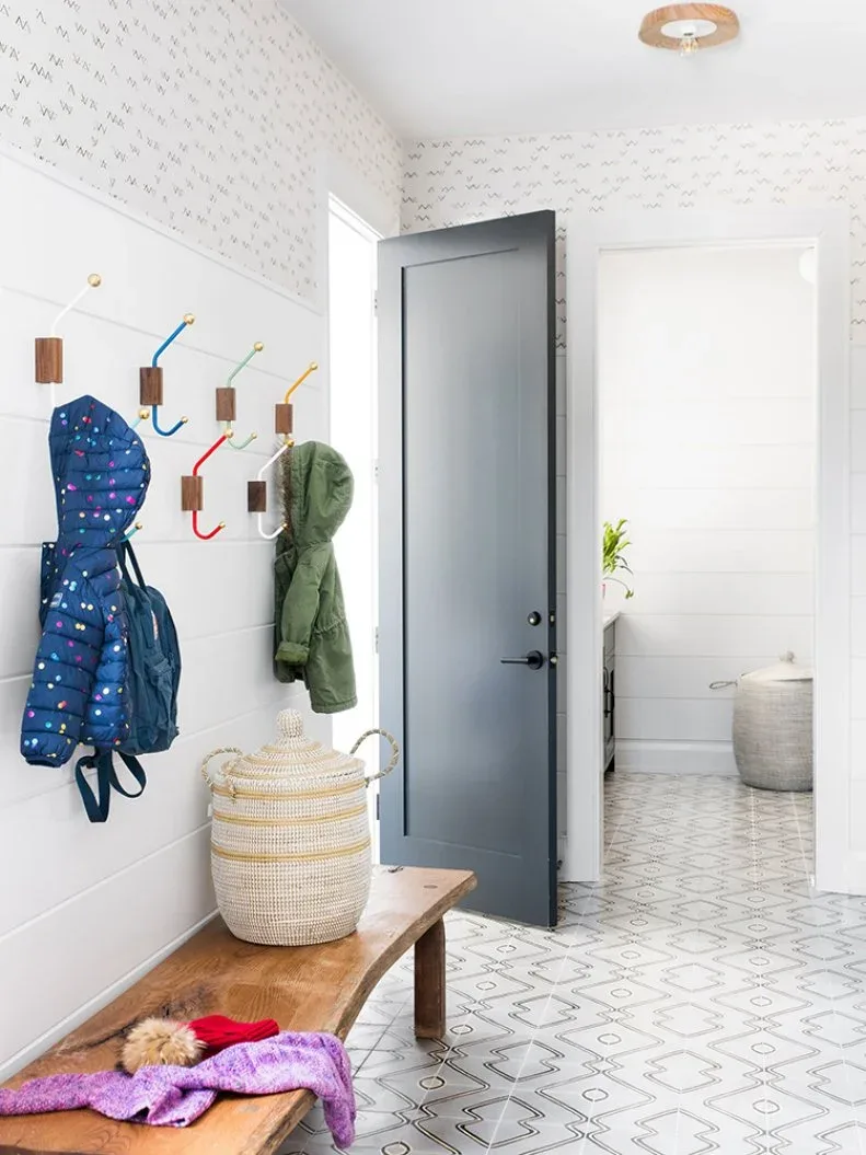 Light mudroom with wooden bench, colorful wall hooks, woven lidded basket, small patterned walls and dark gray door.