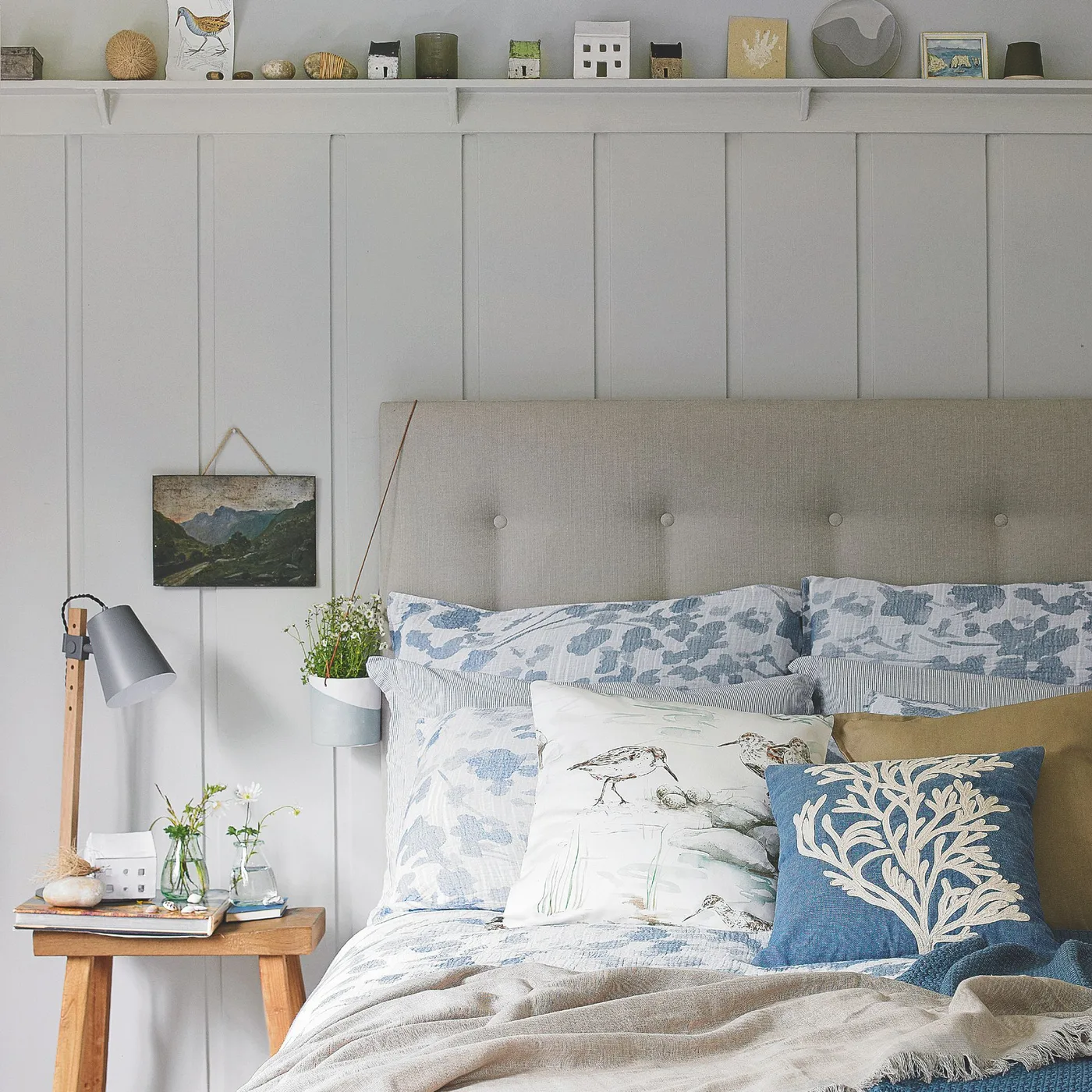 Bedroom with a gray upholstered headboard, pale wall paneling, blue botanical bedding, nautical cushions and a wooden bedside stool.