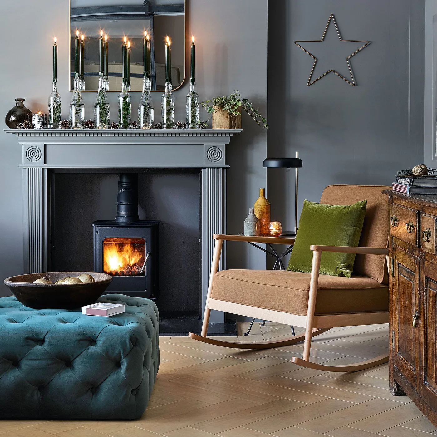 A pale wooden rocking chair sits beside a teal tufted ottoman and dark paneled fireplace wall